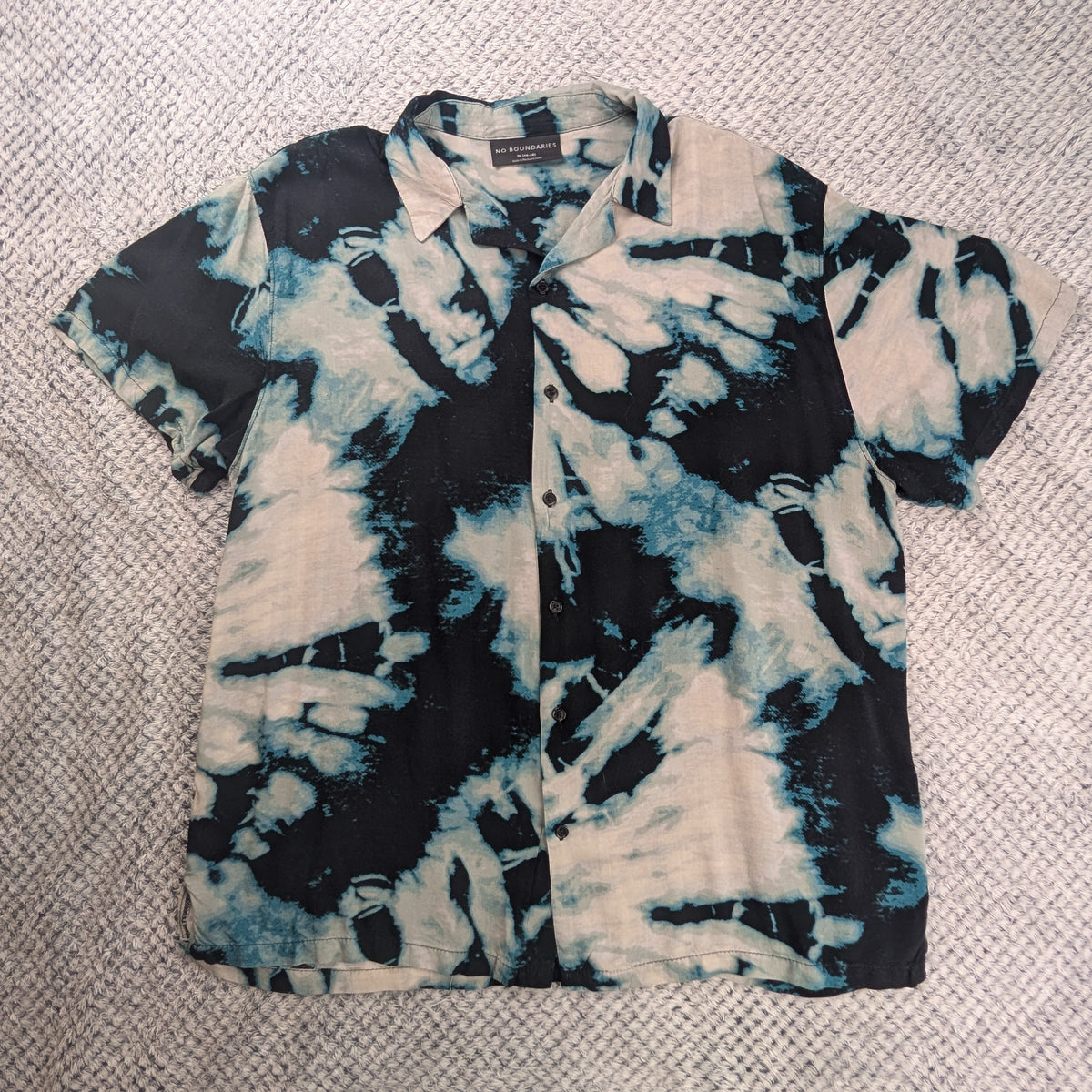 A short-sleeve button-up shirt with a black, white, and blue tie-dye pattern, laid flat on a textured white blanket.