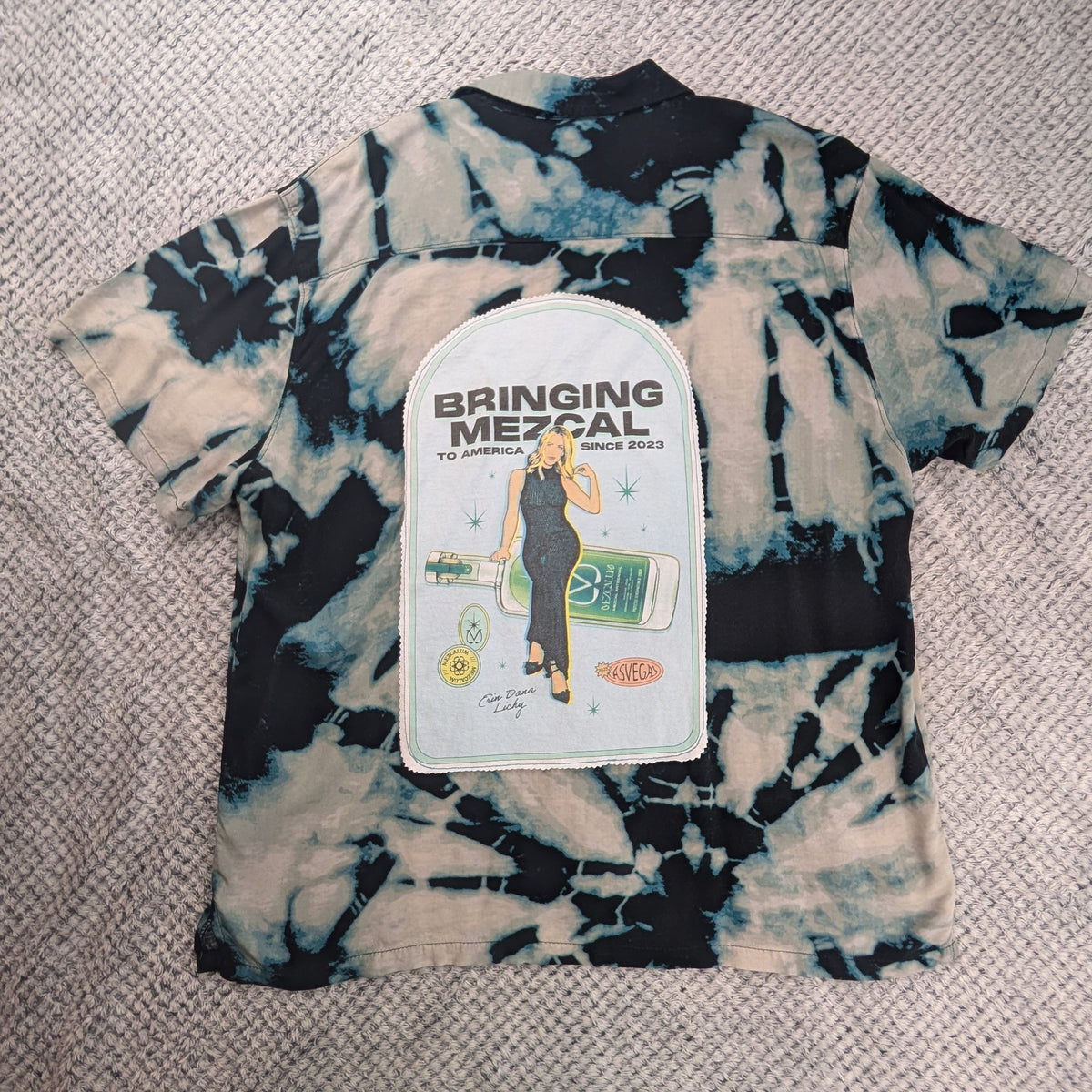 A tie-dye t-shirt with a black, white, and blue pattern, featuring a white graphic patch on the front that reads 'BRINGING THE MEZCAL'. The shirt is laid flat on a textured beige rug.