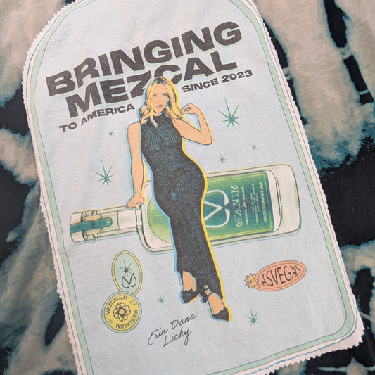 A rectangular fabric patch with a scalloped border featuring a cartoon illustration of a woman holding a green bottle, with the text 'BRINGING MEZCAL TO AMERICA' and 'BRINGING MEZCAL SINCE 2023', placed on a black and white tie-dye surface.