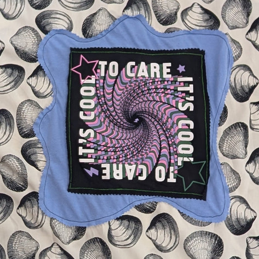 A short-sleeve t-shirt with a black and white seashell pattern, laid flat on a gray knit blanket. The center features a graphic patch with a purple spiral and the text 'TO CARE IT'S GOOD TO CARE'.