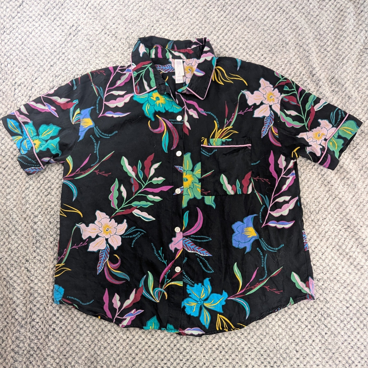 A short-sleeve button-up shirt with a vibrant multicolor floral print on a black background, laid flat on a textured cream-colored blanket.