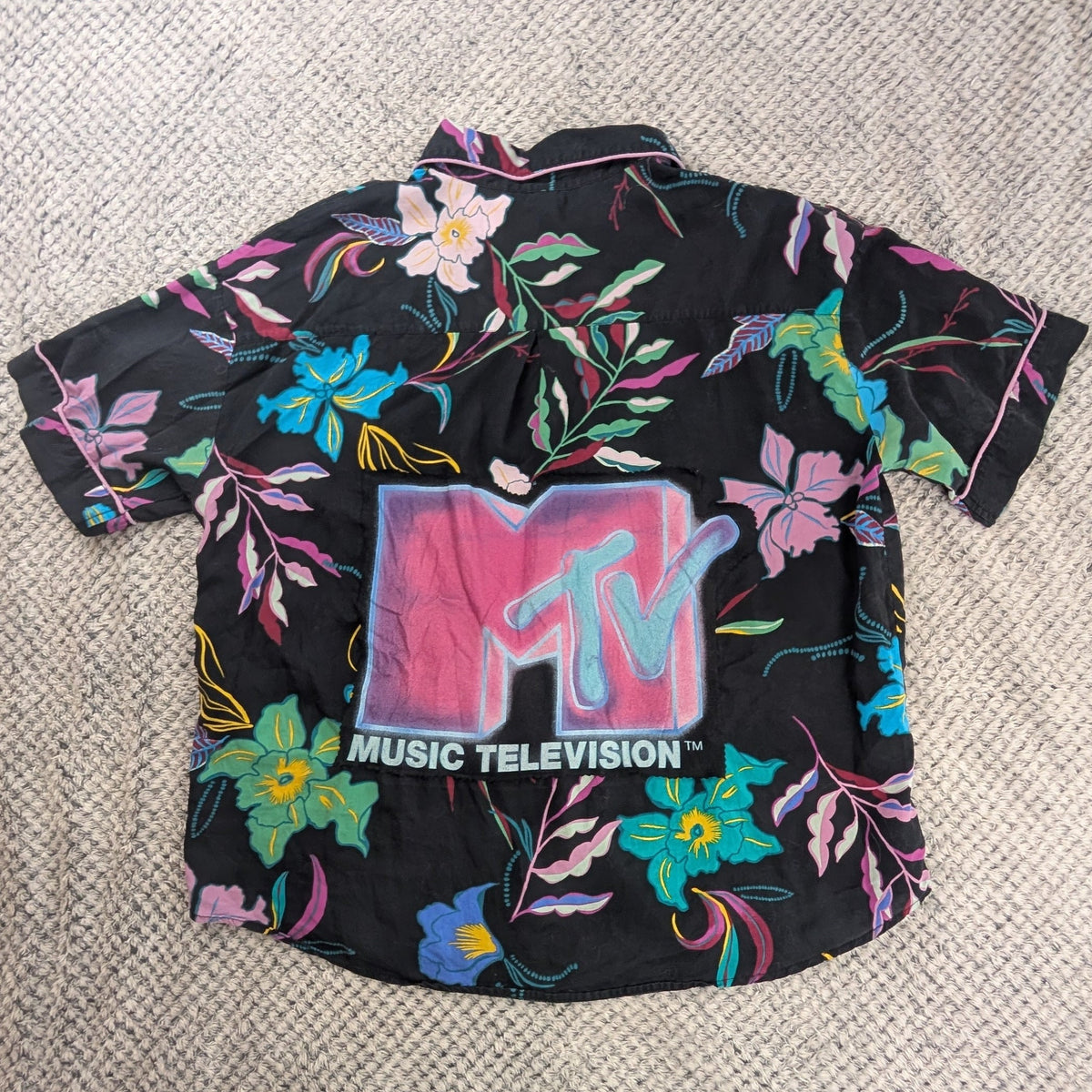 A black short-sleeve button-up shirt with a colorful floral print and a large MTV Music Television logo on the front, laid flat on a textured white blanket with a person's feet visible at the bottom edge.