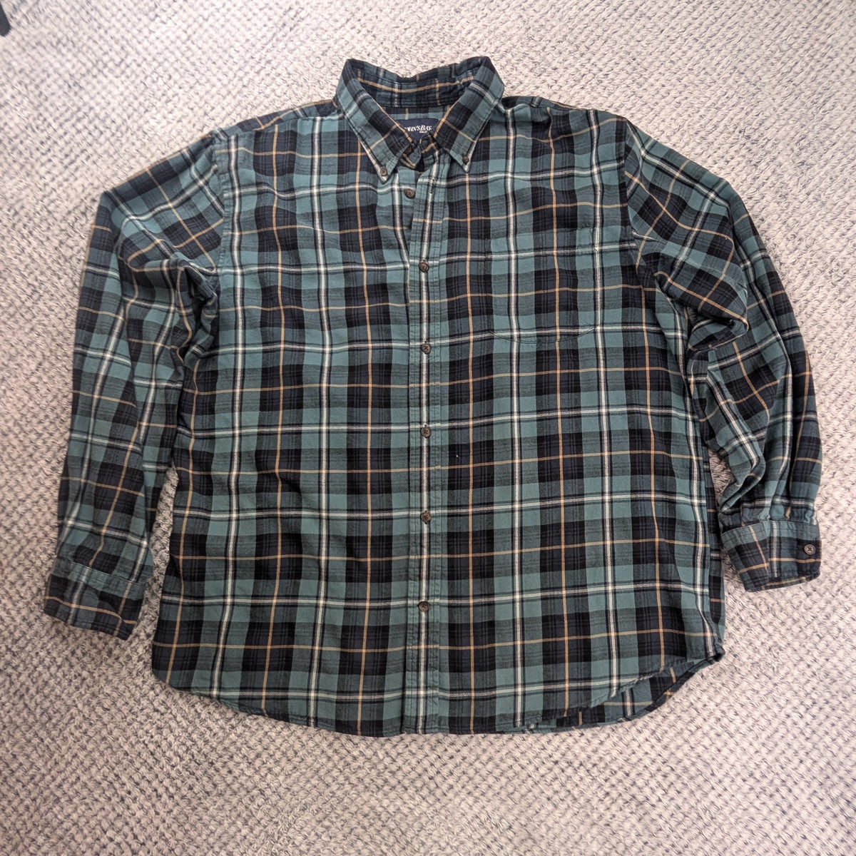 A long-sleeve plaid button-up shirt in green, black, and white, laid flat on a textured white blanket with carpet visible around the edges.