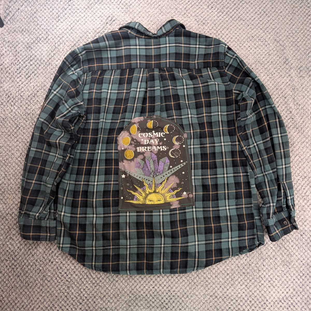 A green and black plaid long-sleeve button-up shirt laid flat on a textured white blanket, featuring a colorful graphic print on the back with the words 'Sugared Love Affair' and 'Love Never Dies'.