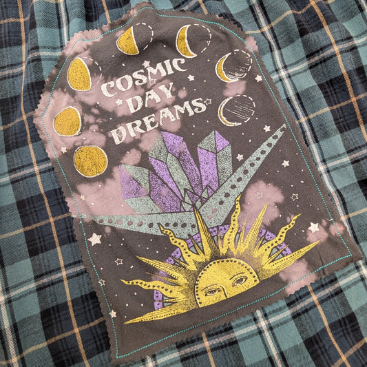 A close-up of a plaid garment, likely pajama pants or a skirt, featuring a decorative patch that reads 'COSMIC DAY DREAMS' with a purple crystal, yellow sun, and moon phases on a dark gray background.