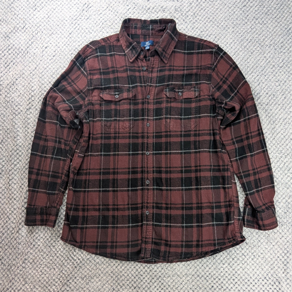A long-sleeve plaid button-up shirt in red and black laid flat on a textured white blanket.