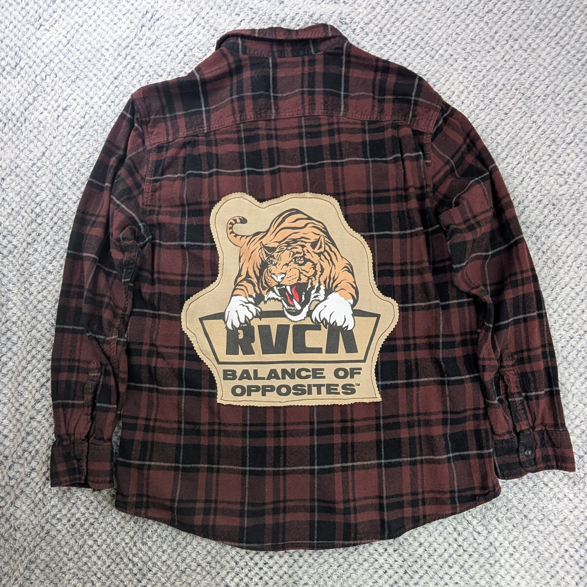 A red and black plaid flannel shirt jacket laid flat on a textured white rug, featuring a large graphic patch on the back with a tiger and the text 'RVCA BALANCE OF OPPOSITES'.