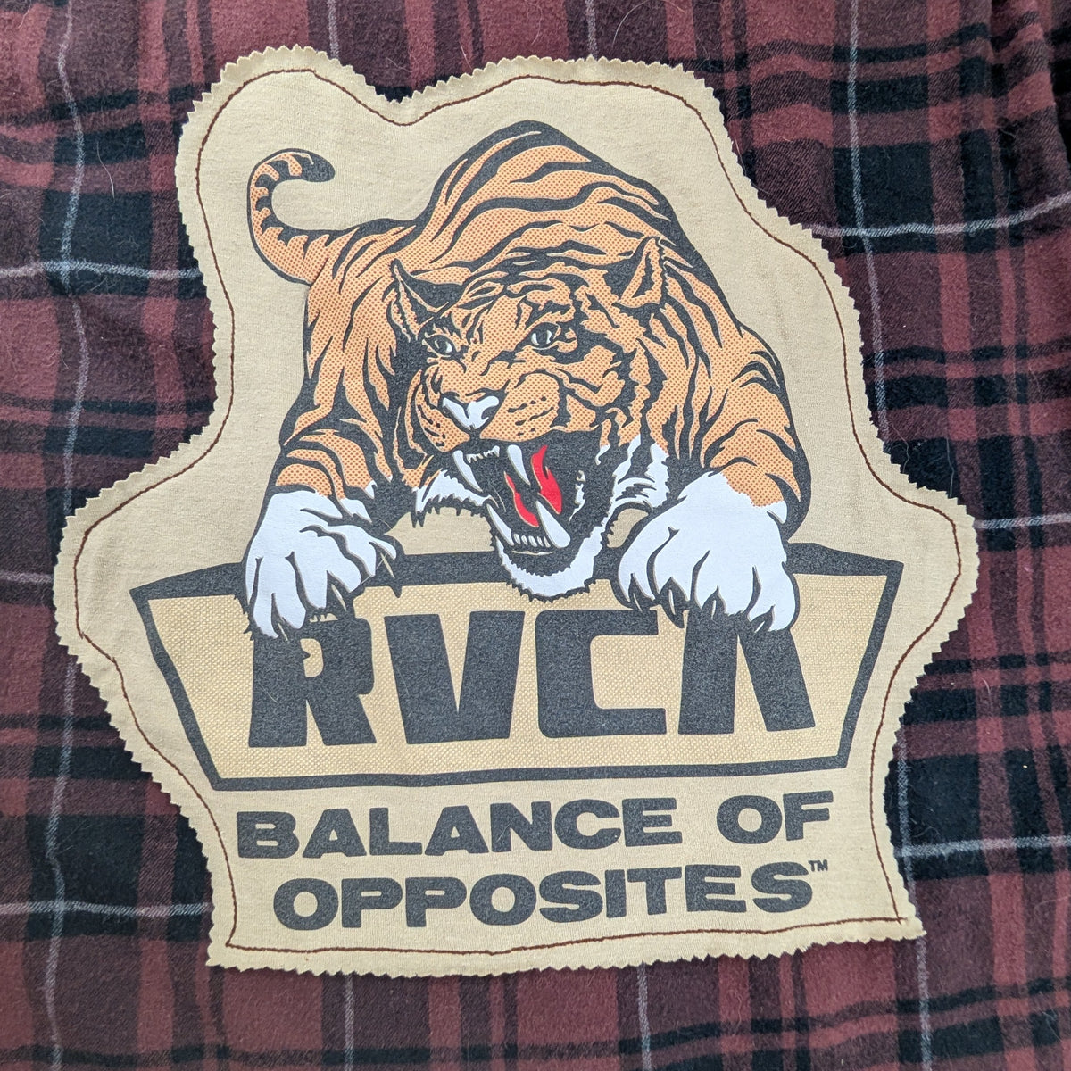 A close-up of a red and black plaid shirt featuring a large embroidered patch with a tiger graphic and the text 'RVCA' and 'BALANCE OF OPPOSITES'.