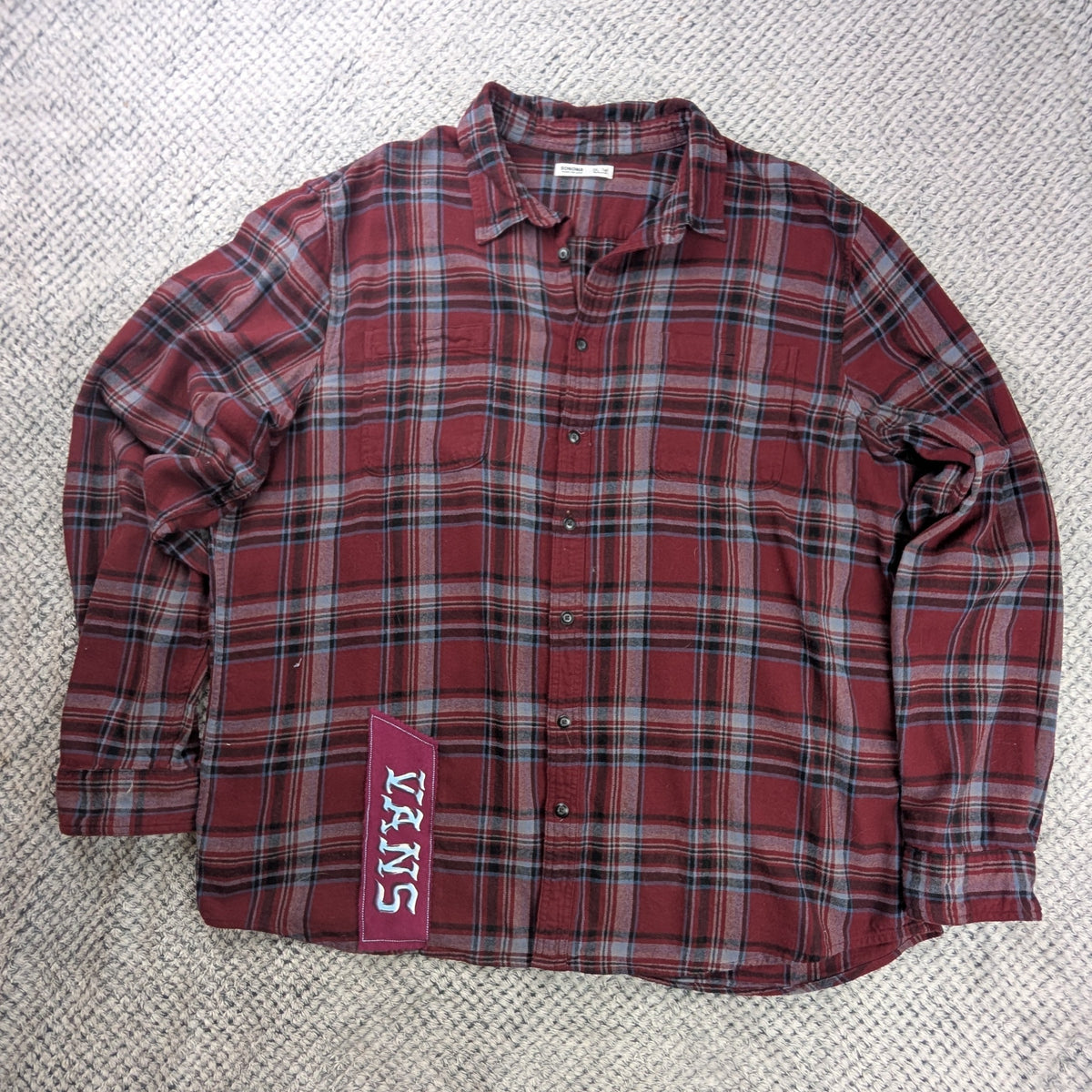 A red and black plaid long-sleeve button-up shirt laid flat on a white textured blanket, with a visible 'Vans' tag on the lower left side.