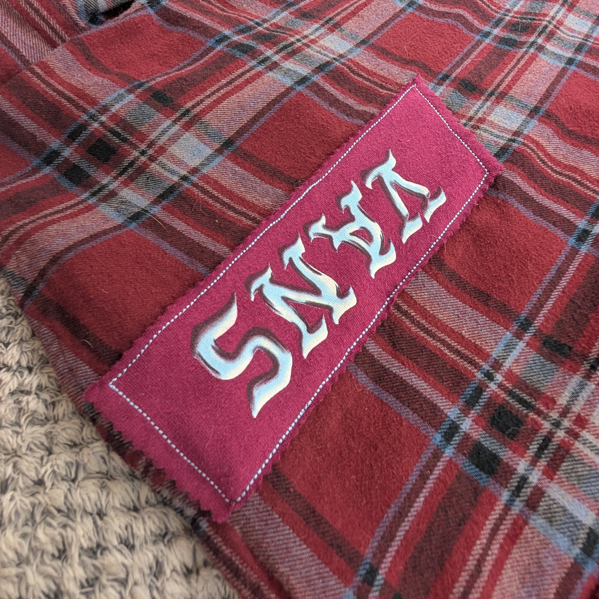 A close-up of a red and black plaid flannel shirt with a pink embroidered patch on the sleeve that reads 'SNEZ' in white gothic lettering, laid on a textured beige blanket.