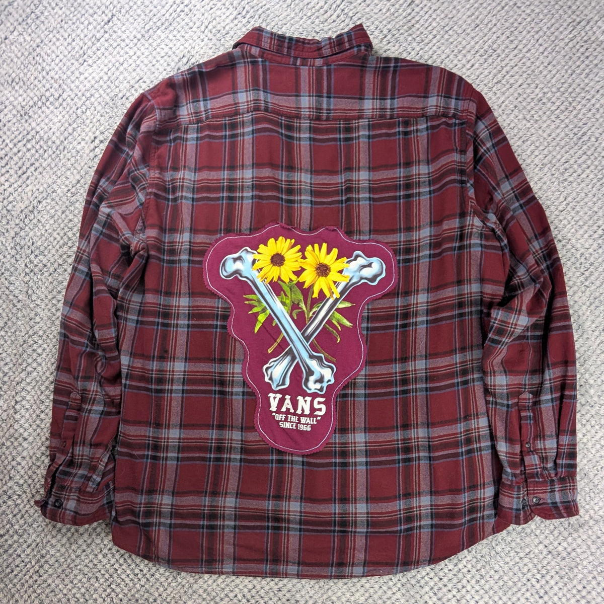 A red and black plaid flannel shirt laid flat on a white textured blanket, featuring a large back graphic with crossed bones, yellow sunflowers, and the text 'VANS' and 'EST. 2015'.