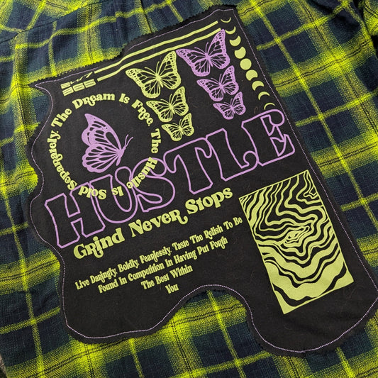 A green and black plaid flannel shirt laid flat on a carpeted floor, featuring a large black graphic patch on the front with purple and yellow text and butterfly illustrations.