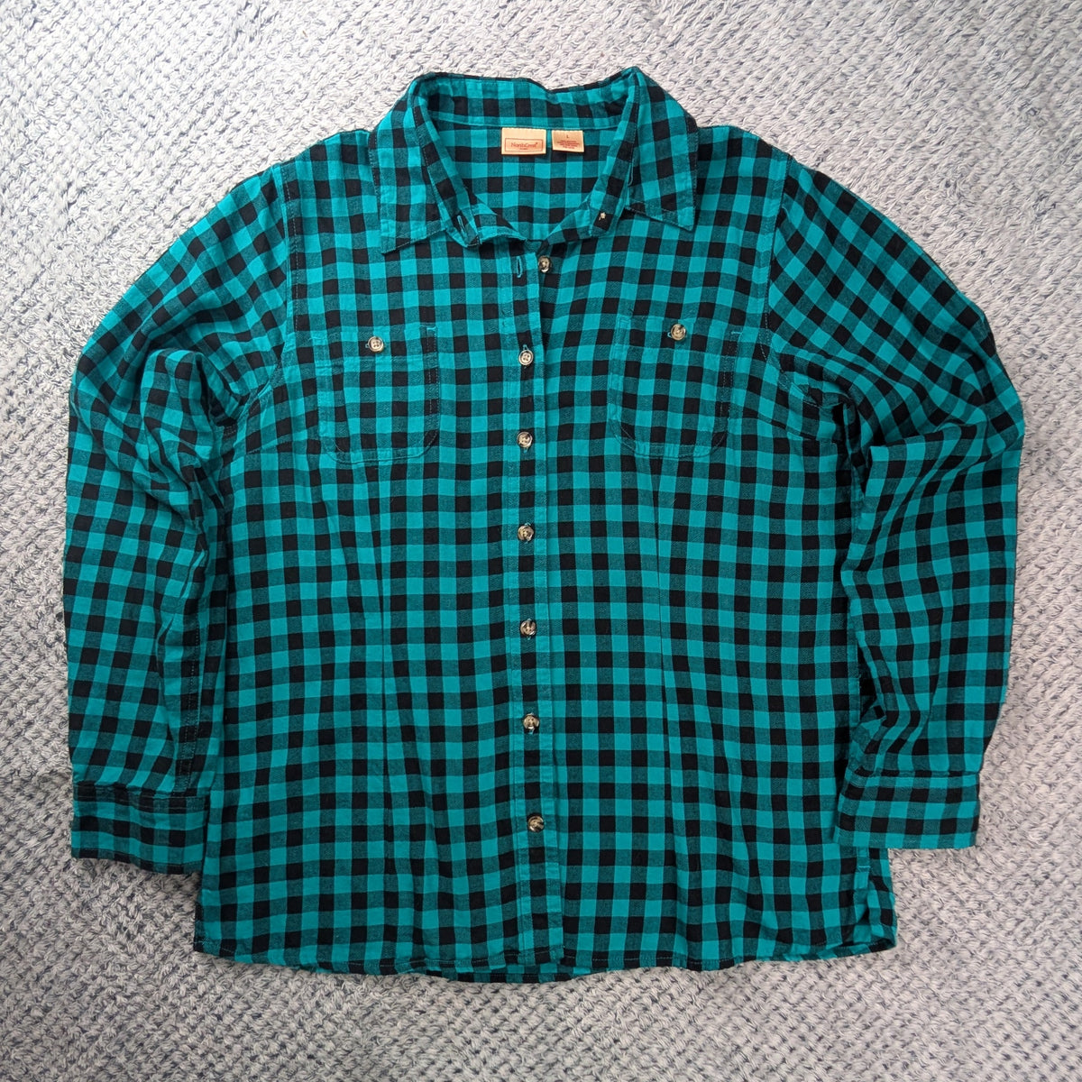 A teal and black checkered long-sleeve flannel shirt laid flat on a gray textured blanket, featuring a large graphic print of a cartoon car on a white cloud shape on the back.