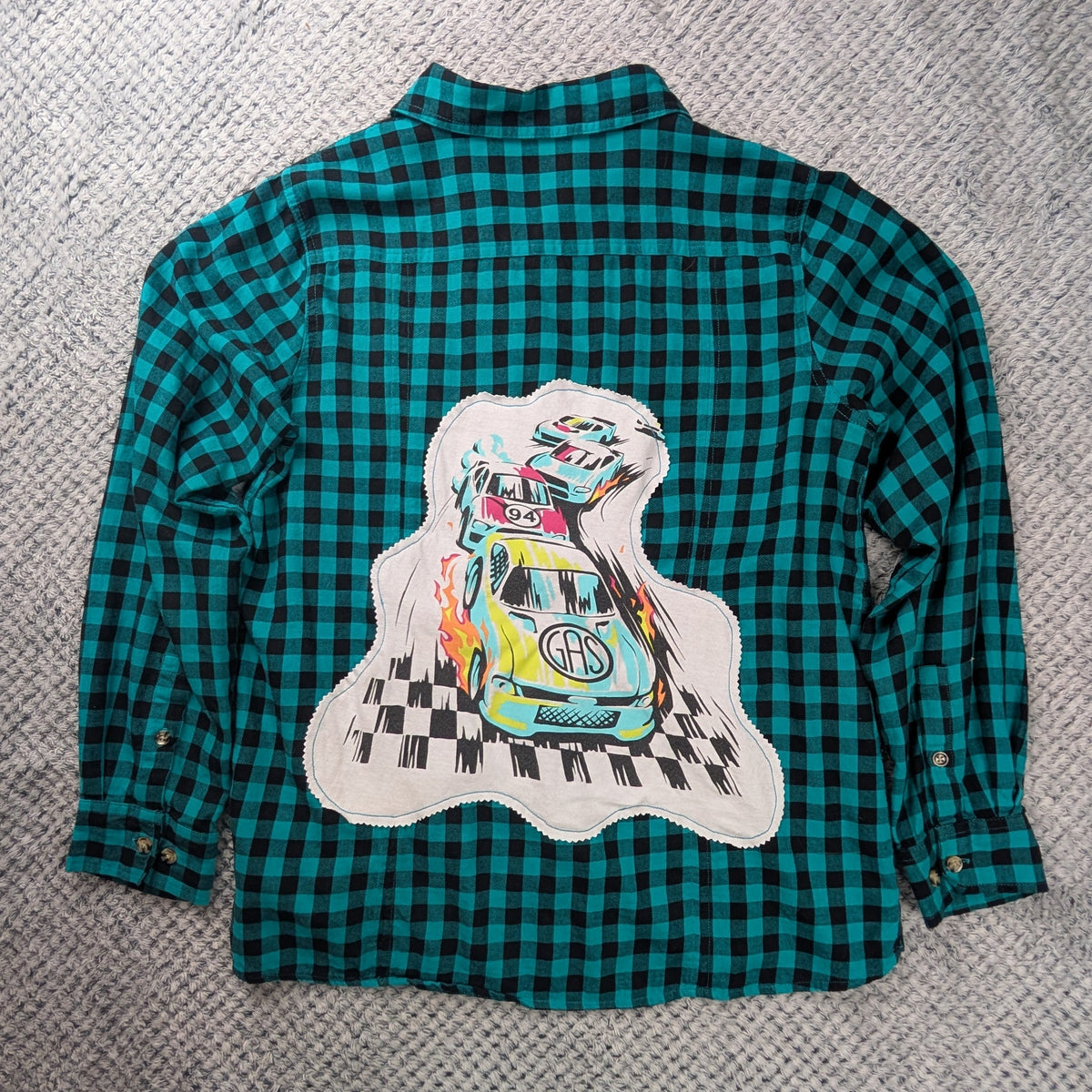 A teal and black checkered long-sleeve flannel shirt laid flat on a gray textured blanket, featuring a large graphic print of a cartoon car on a white cloud shape on the back.