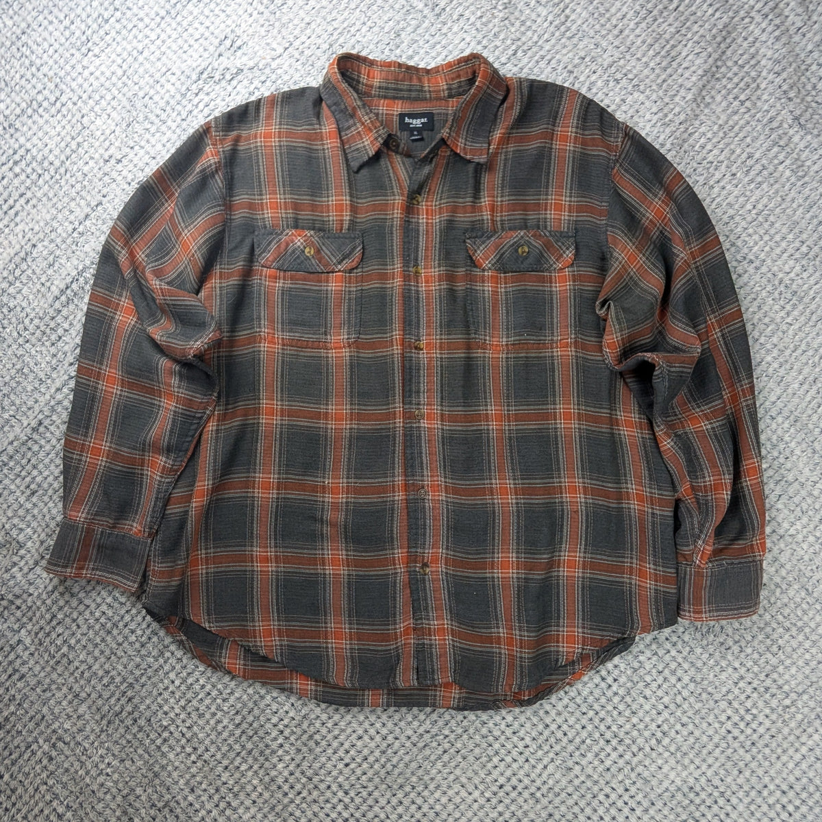 A long-sleeve plaid button-up shirt laid flat on a textured light gray blanket. The shirt features a dark gray and black plaid pattern with orange-brown lines.