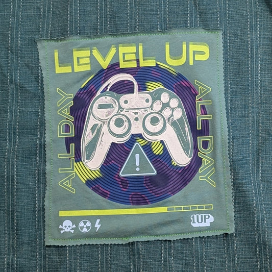 A dark teal-green short-sleeve button-up shirt with vertical pinstripes, featuring a colorful retro video game 'Level Up' graphic patch on the back, laid flat on a textured gray blanket.