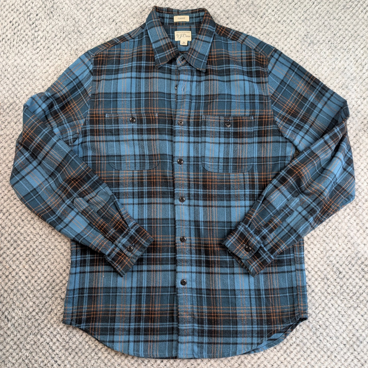 A long-sleeve button-up flannel shirt in a blue, black, and brown plaid pattern, laid flat on a textured gray woven rug.