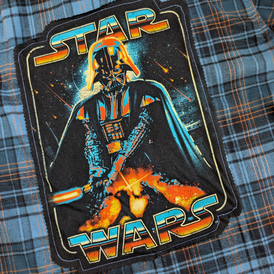 A close-up of a Star Wars graphic patch featuring Darth Vader with a lightsaber, placed on a blue and orange plaid fabric background.