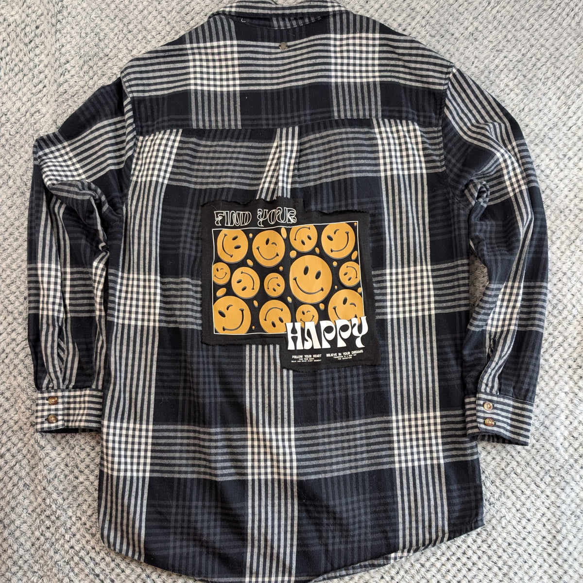 A black and white plaid flannel shirt laid flat on a textured gray carpet, featuring a large graphic patch on the back with yellow smiley faces and the word 'HAPPY'.