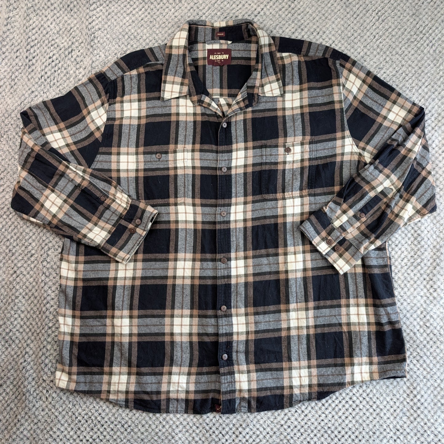 A long-sleeve plaid button-up shirt laid flat on a textured gray blanket. The shirt features a dark navy, gray, and beige plaid pattern with a small embroidered logo on the left chest.