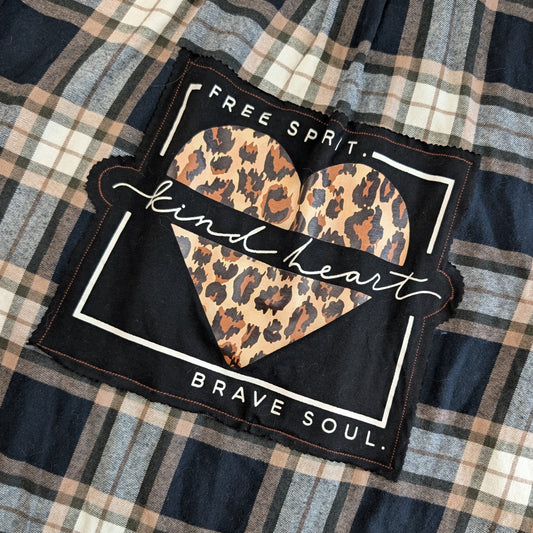 A square black fabric patch with a leopard-print heart in the center, bordered in white and overlaid with a white script banner reading 'kind heart.' The patch is placed on a black, gray, and beige background.