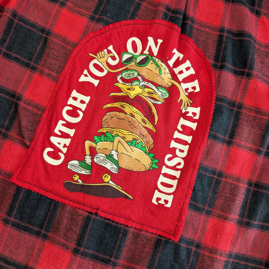 A close-up of a red patchwork fabric featuring a large red applique patch with a cartoon taco character wearing sunglasses and the text 'CATCH YOU ON THE FLIPSIDE,' laid over a red and black plaid background.