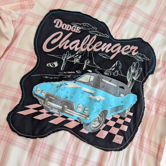 A close-up of a black fabric patch featuring a distressed light blue Dodge Challenger car, cacti, mountains, and the words 'Dodge Challenger' in pink and white, laid flat on a pink and white plaid fabric background.