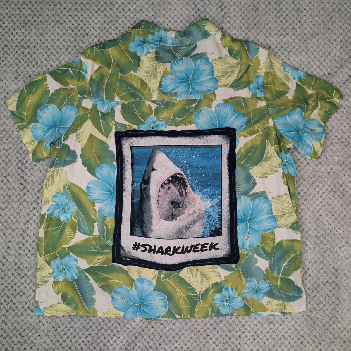 A short-sleeve Hawaiian-style shirt laid flat on a gray textured blanket. The shirt features a green and blue tropical floral print with a large square graphic on the front showing a shark in water and the text '#SHARKWEEK'.