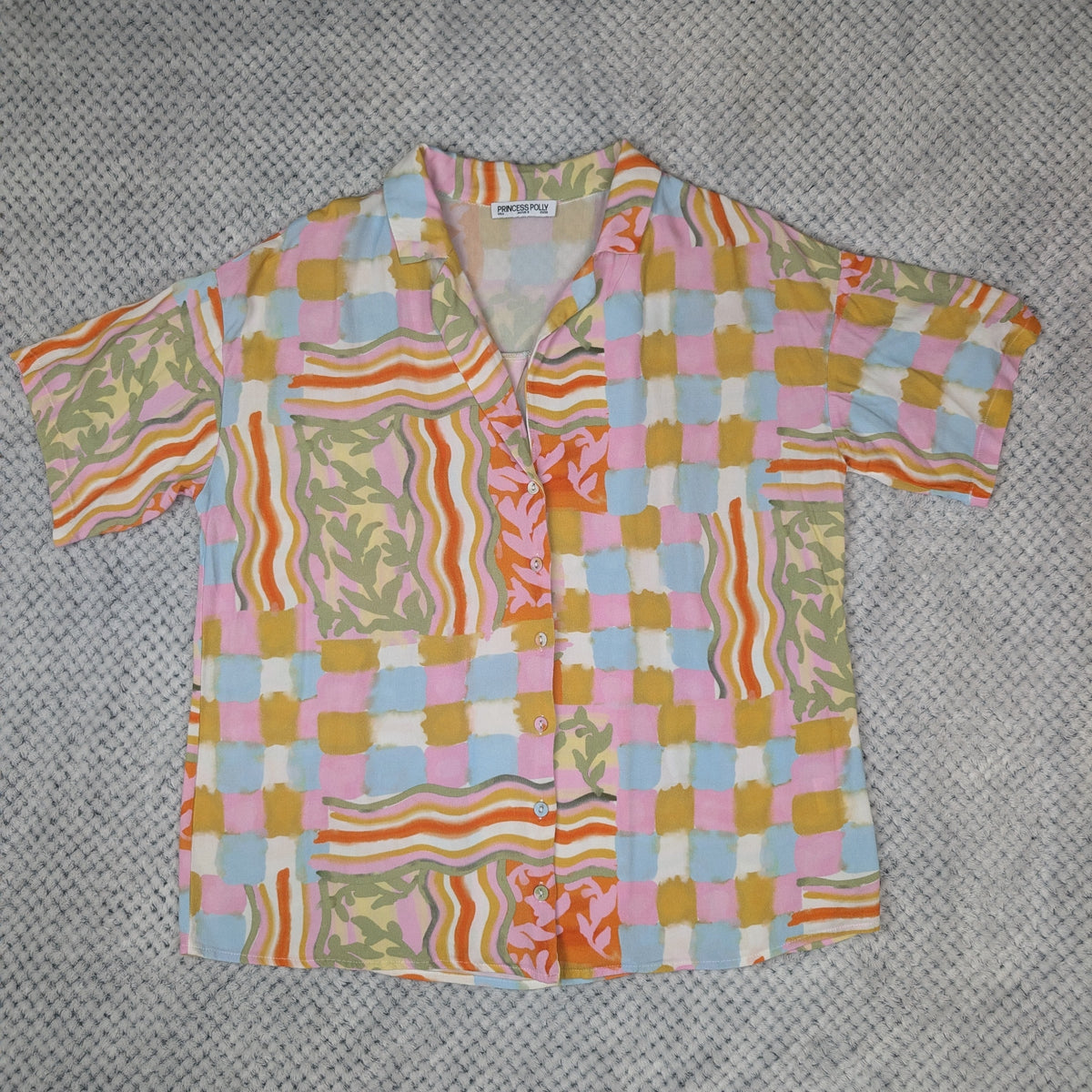 A short-sleeve button-up shirt with a retro multicolor patchwork pattern, laid flat on a gray textured fabric background.