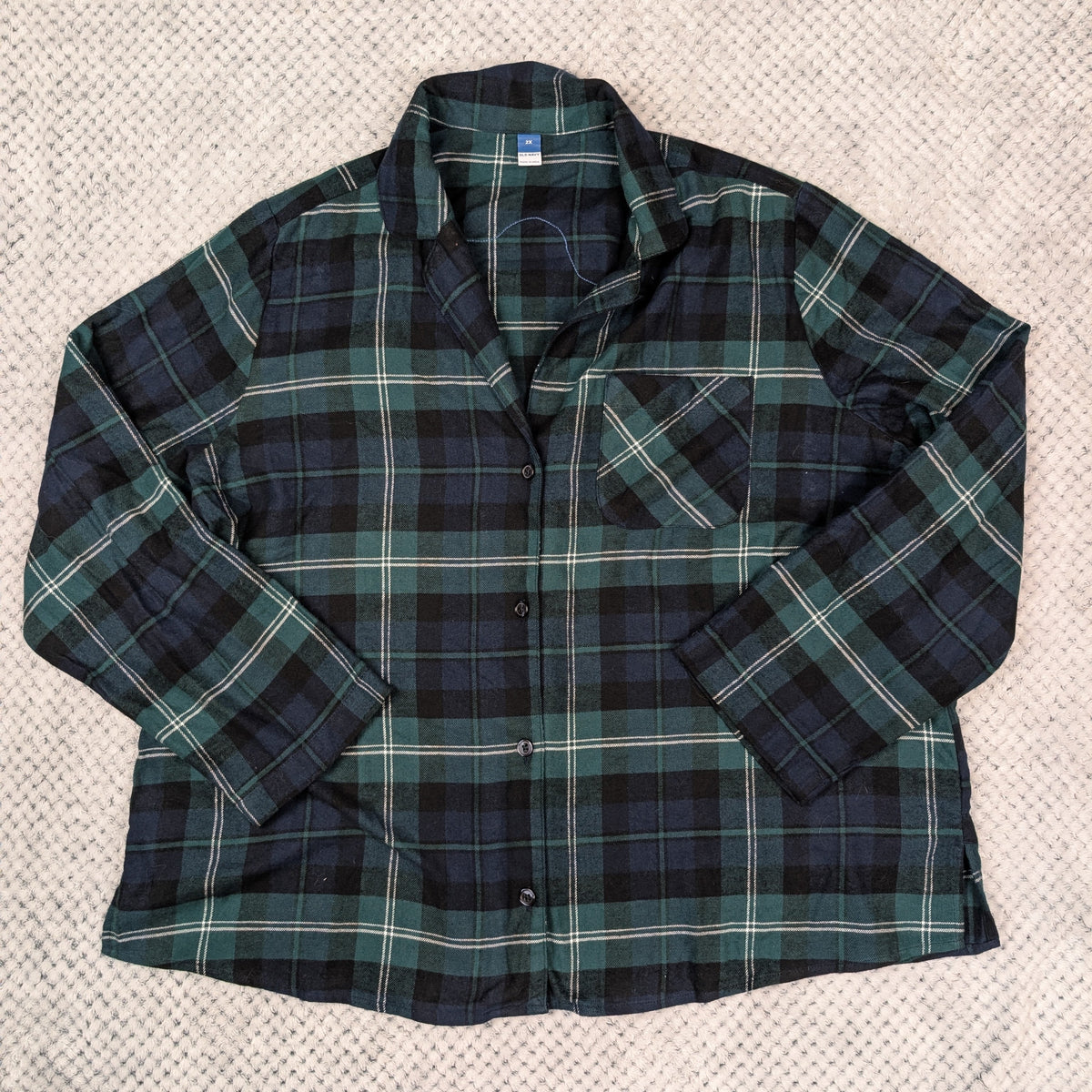 A men's long-sleeve plaid button-up shirt in dark green, navy, and black, laid flat on a textured beige blanket.
