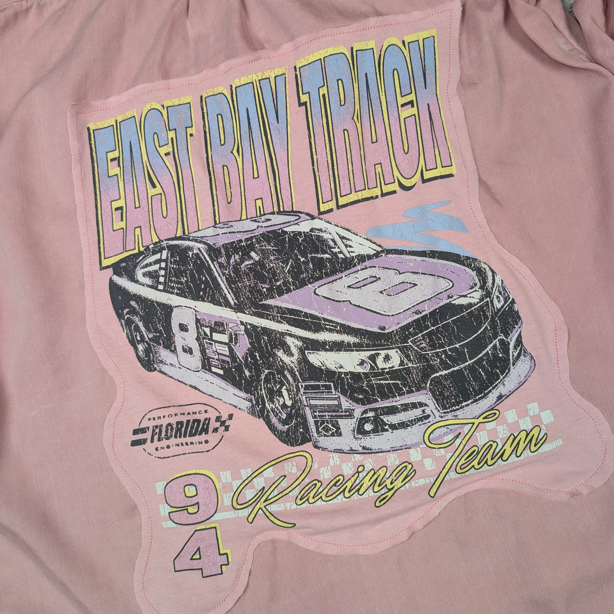A close-up of a dusty pink garment featuring a large graphic print on the front. The print shows a black and purple race car with the number 9, the words 'EAST BAY TRACK' in blue and yellow, 'Florida Racing Team' in green script, and the number 94.