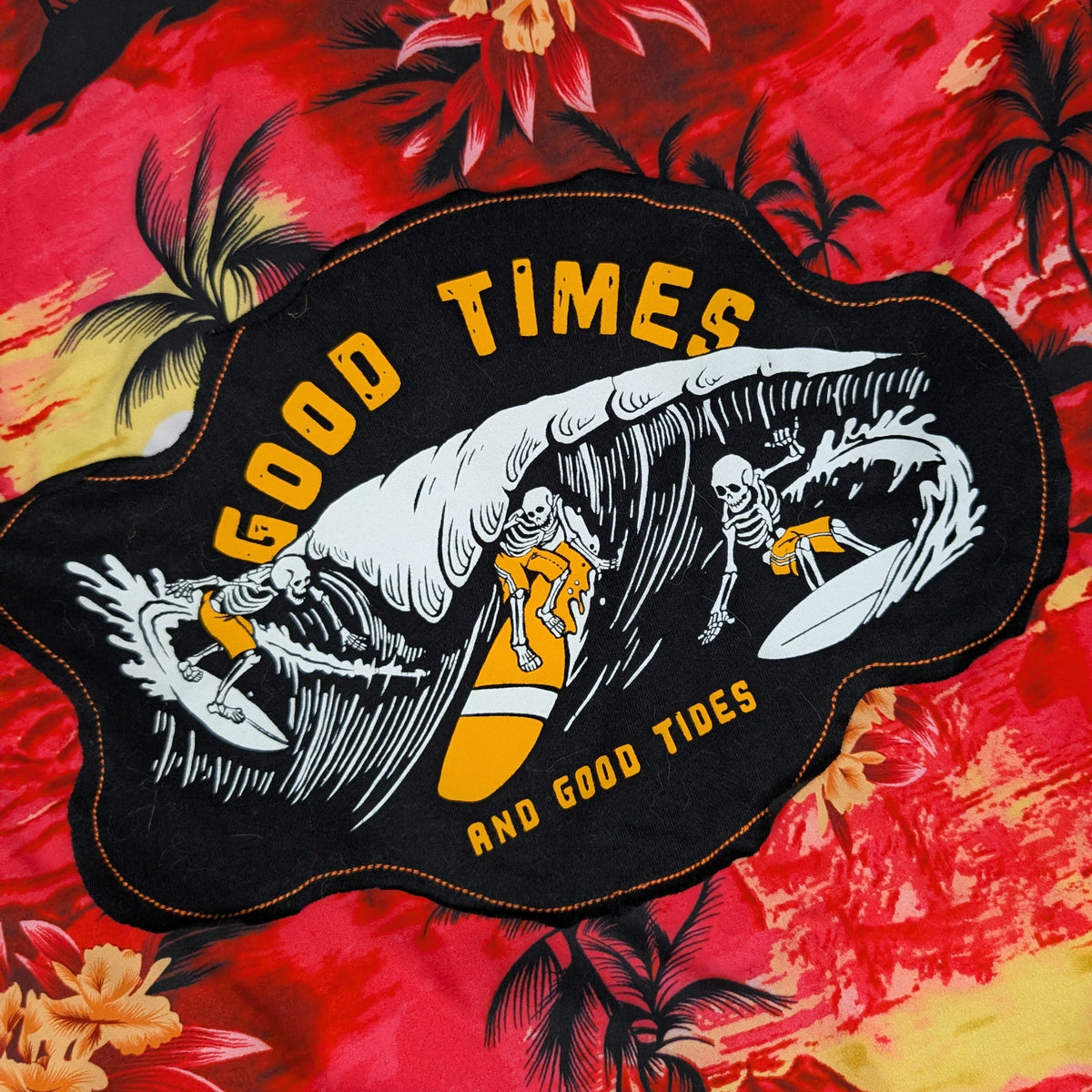 A close-up of a red tropical floral and palm tree print fabric with a large black embroidered patch featuring a surfer wave graphic and the words 'GOOD TIMES AND GOOD TIDES'.