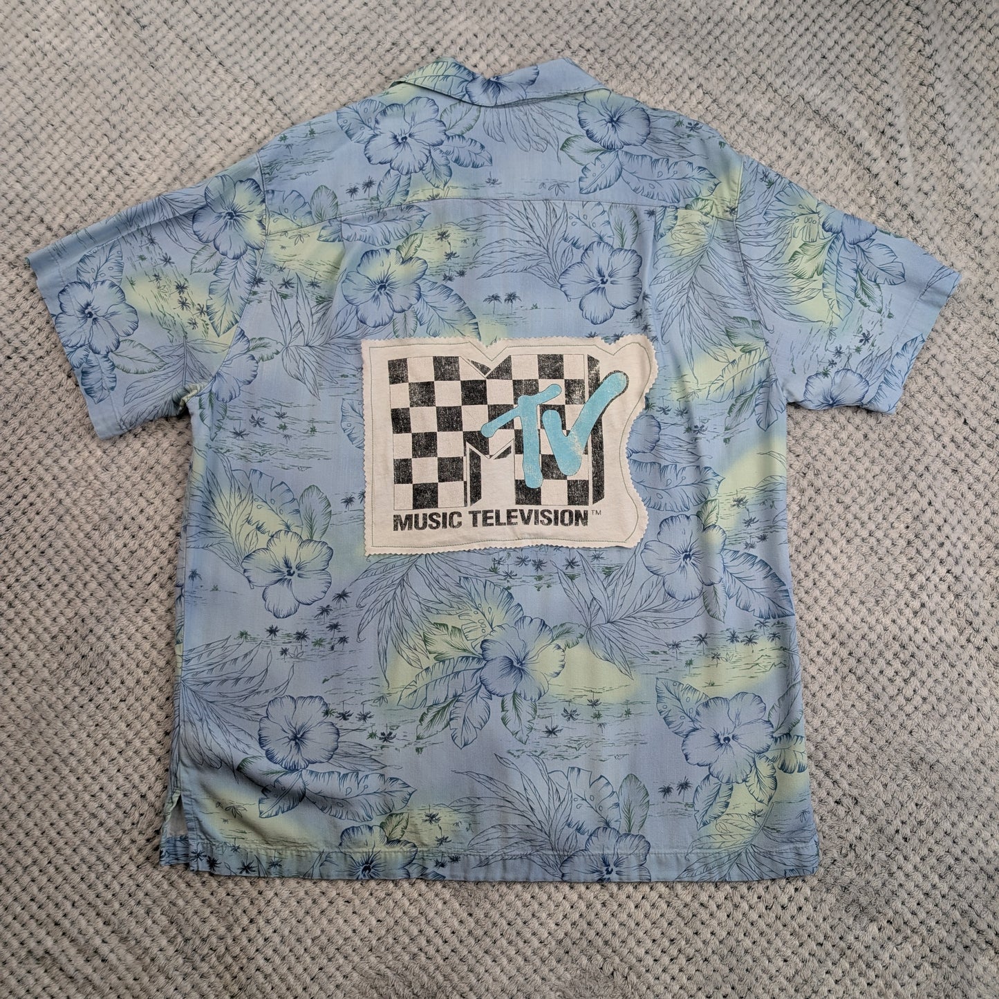 A light blue short-sleeve button-up shirt with a tropical floral print in pale yellow and green, featuring a large MTV Music Television patch on the back, laid flat on a textured gray blanket.