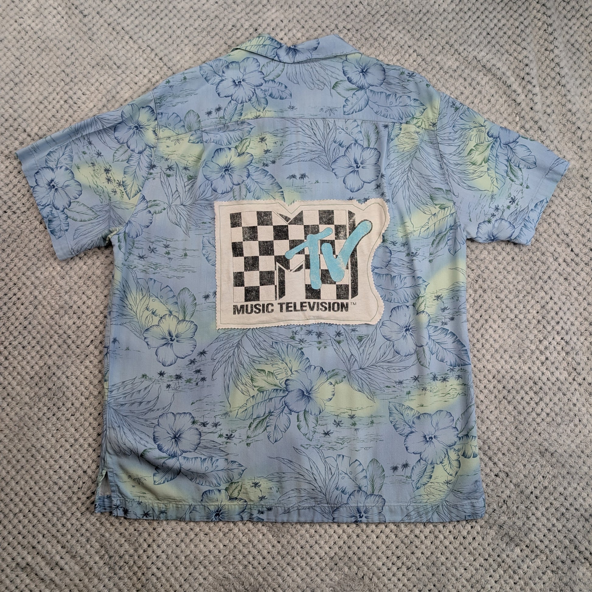 A light blue short-sleeve button-up shirt with a tropical floral print in pale yellow and green, featuring a large MTV Music Television patch on the back, laid flat on a textured gray blanket.