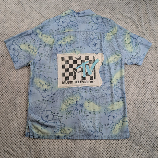 A light blue short-sleeve button-up shirt with a tropical floral print in pale yellow and green, featuring a large MTV Music Television patch on the back, laid flat on a textured gray blanket.