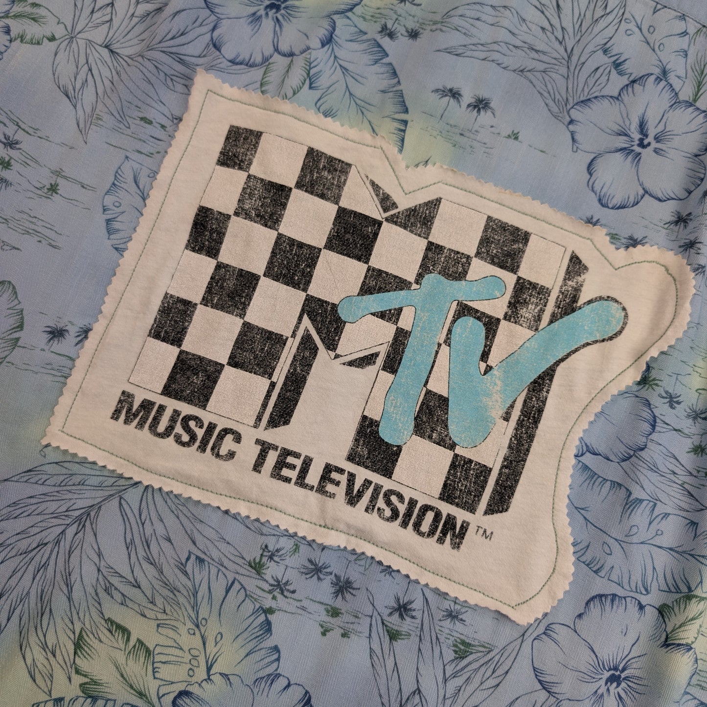 A vintage MTV Music Television embroidered patch with a black and white checkerboard logo and light blue 'TV' lettering, displayed on a blue floral fabric background.