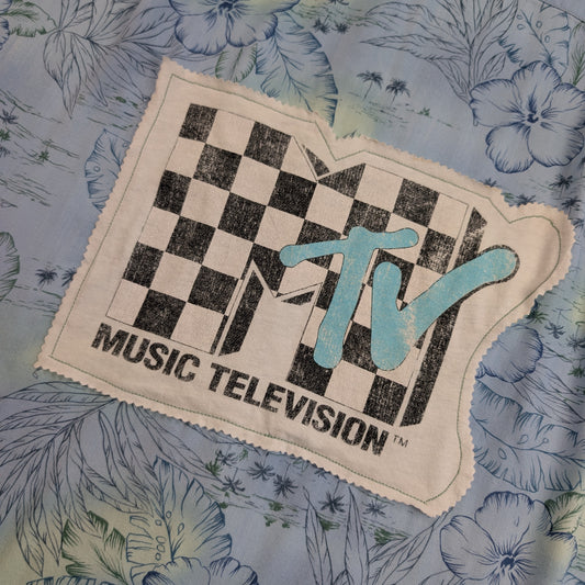 A vintage MTV Music Television embroidered patch with a black and white checkerboard logo and light blue 'TV' lettering, displayed on a blue floral fabric background.
