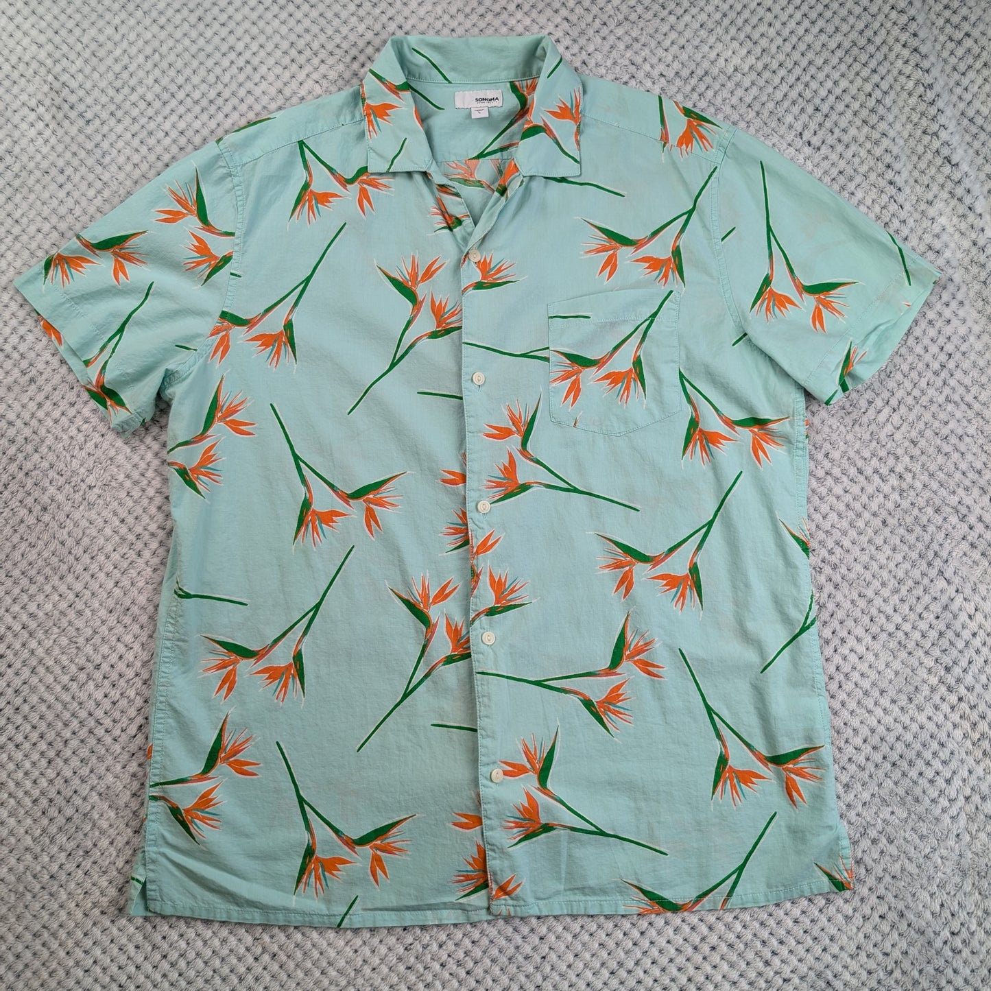A short-sleeve button-up shirt in a light mint green color, featuring an all-over tropical bird of paradise flower print in orange and green, laid flat on a textured gray blanket.