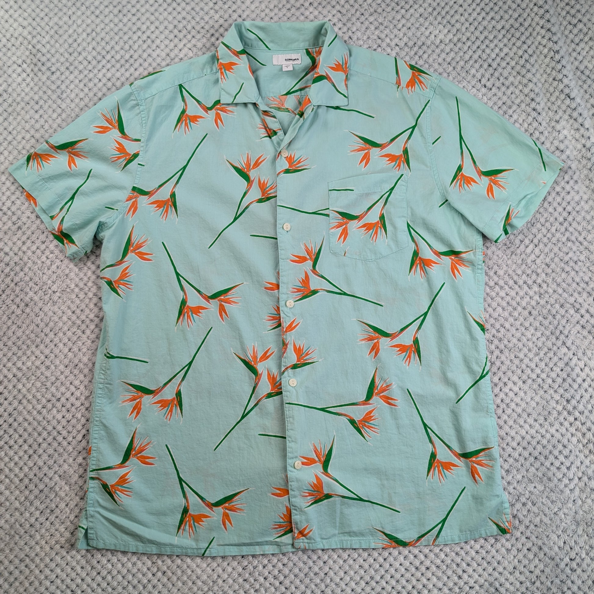 A short-sleeve button-up shirt in a light mint green color, featuring an all-over tropical bird of paradise flower print in orange and green, laid flat on a textured gray blanket.