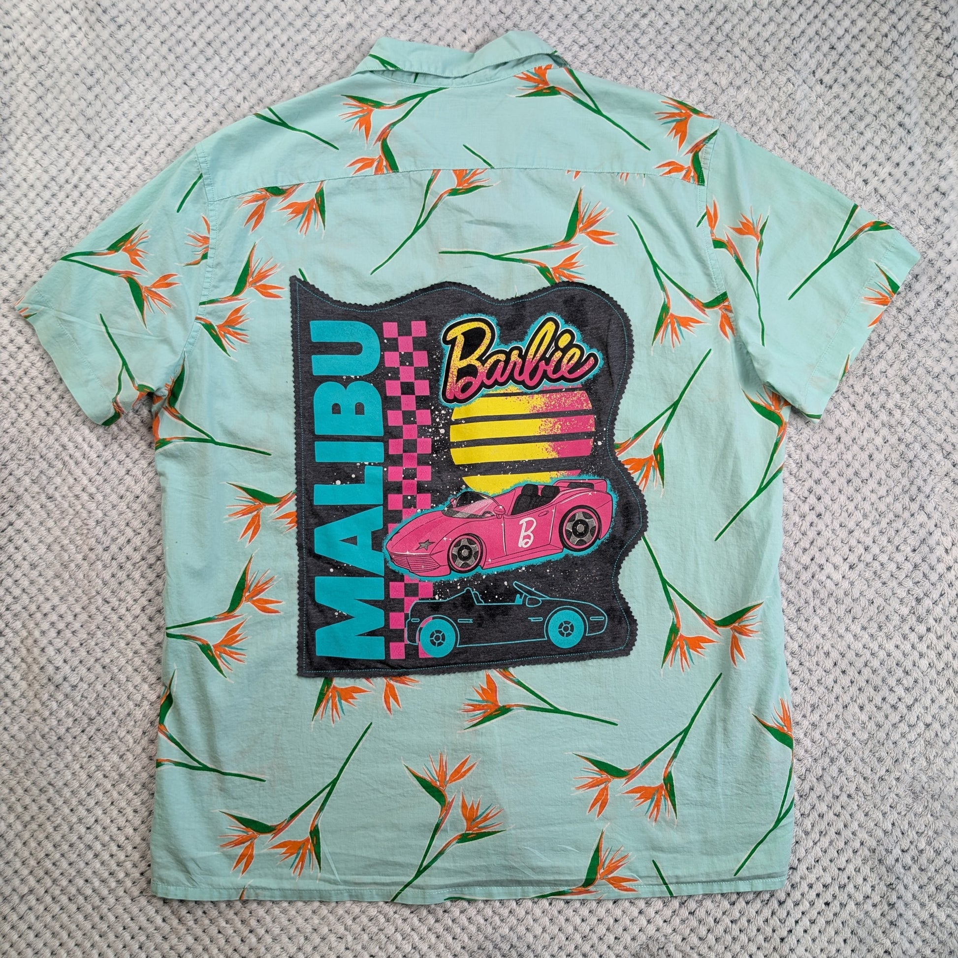 A mint green short-sleeve button-up shirt with an all-over orange and green tropical floral print, featuring a large Barbie Malibu graphic on the back.