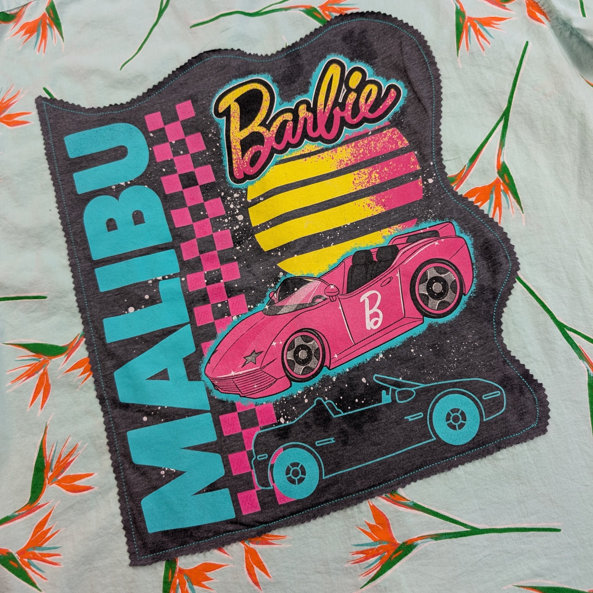 A close-up of a Barbie-themed fabric patch laid flat on a light-colored floral surface. The patch is dark gray with a scalloped edge and features the word 'MALIBU' in large turquoise letters, a pink and yellow striped sun, a pink race car, a black race car, and the Barbie logo in pink and yellow.