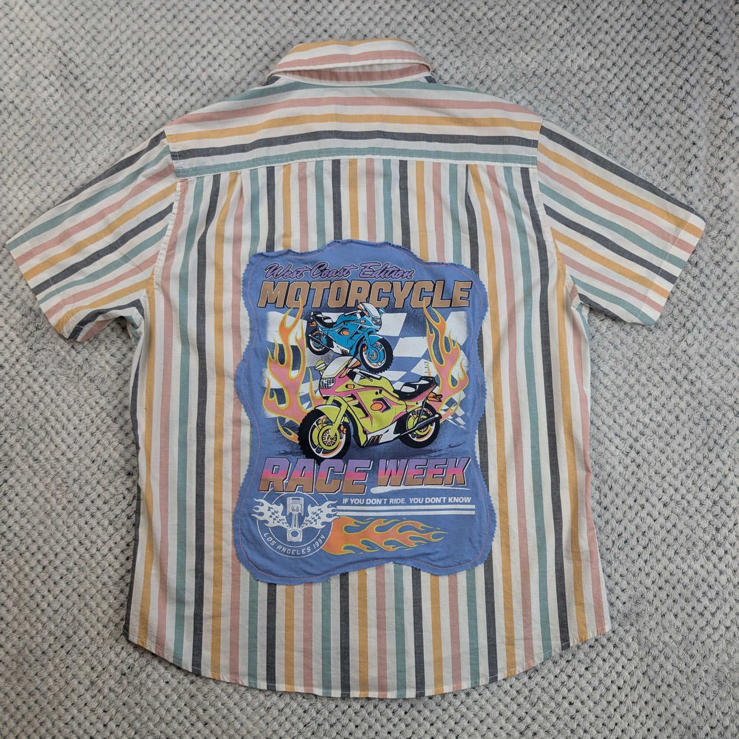 A short-sleeve button-up shirt with vertical stripes in beige, orange, green, and gray, featuring a large blue graphic print on the back with illustrations of motorcycles, flames, and the text 'MOTORCYCLE RACE WEEK'.