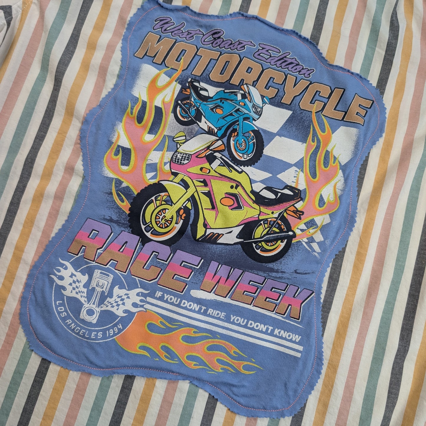 A blue fabric patch with a colorful graphic of two motorcycles and flames, reading 'Ride Like a Dream Motorcycle Race Week', laid flat on a striped fabric background.