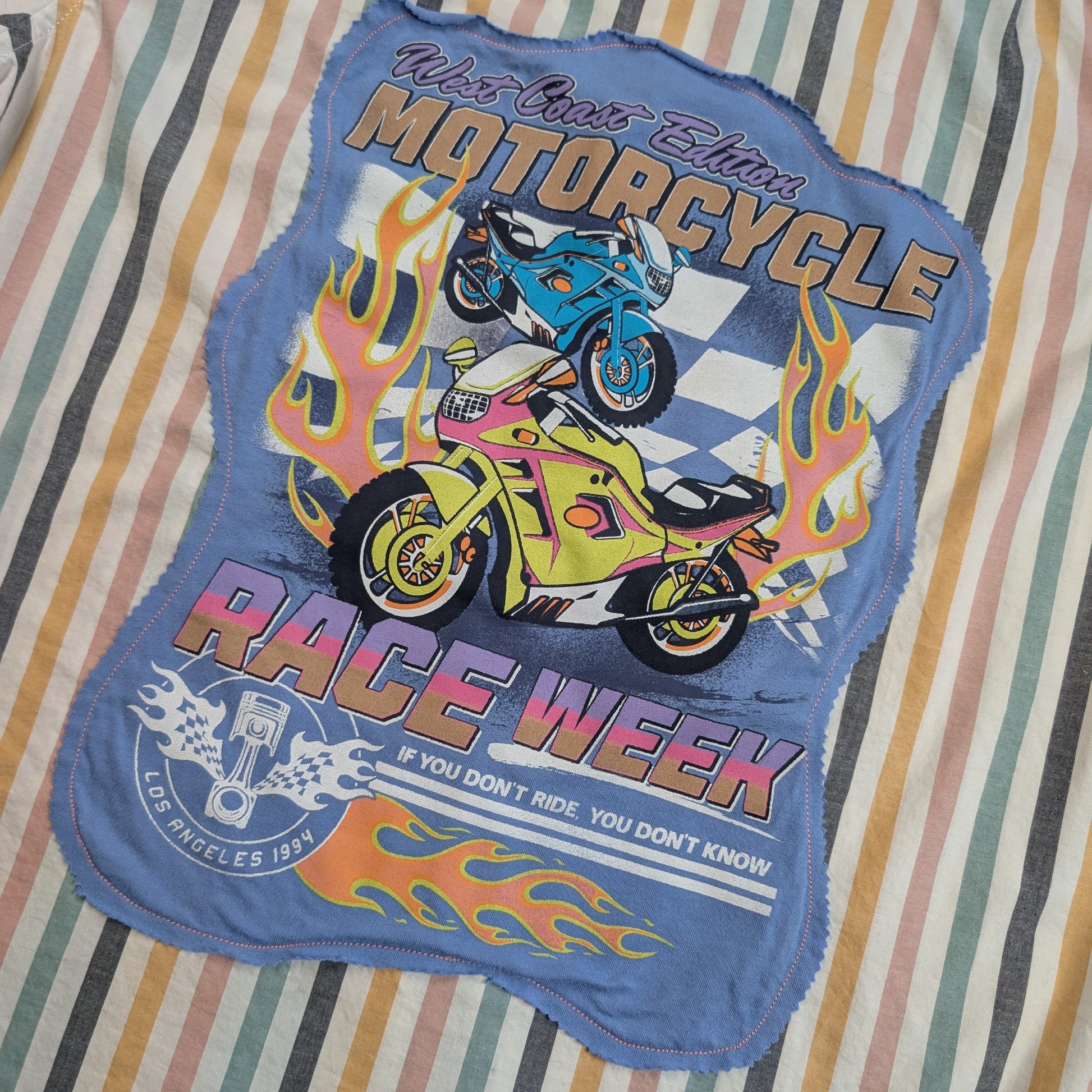 A blue fabric patch with a colorful graphic of two motorcycles and flames, reading 'Ride Like a Dream Motorcycle Race Week', laid flat on a striped fabric background.