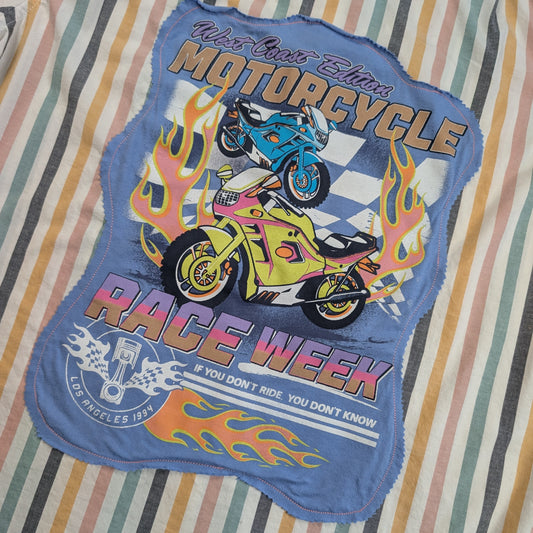 A blue fabric patch with a colorful graphic of two motorcycles and flames, reading 'Ride Like a Dream Motorcycle Race Week', laid flat on a striped fabric background.