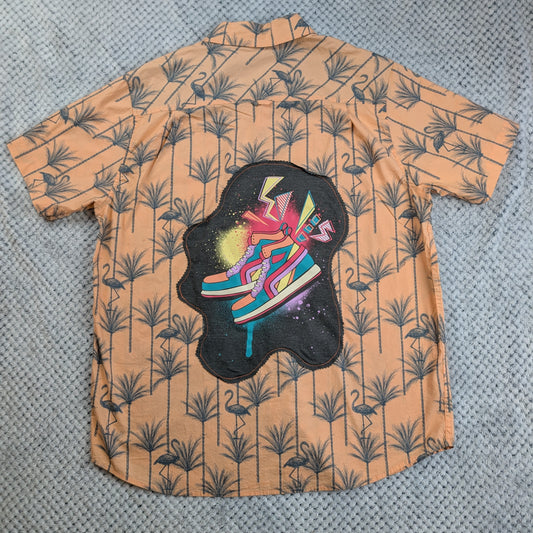 A short-sleeve button-up shirt laid flat on a gray textured surface. The shirt has a peach-orange base with a dark gray palm tree and flamingo pattern, featuring a large central graphic of colorful sneakers on a black cloud-like shape.