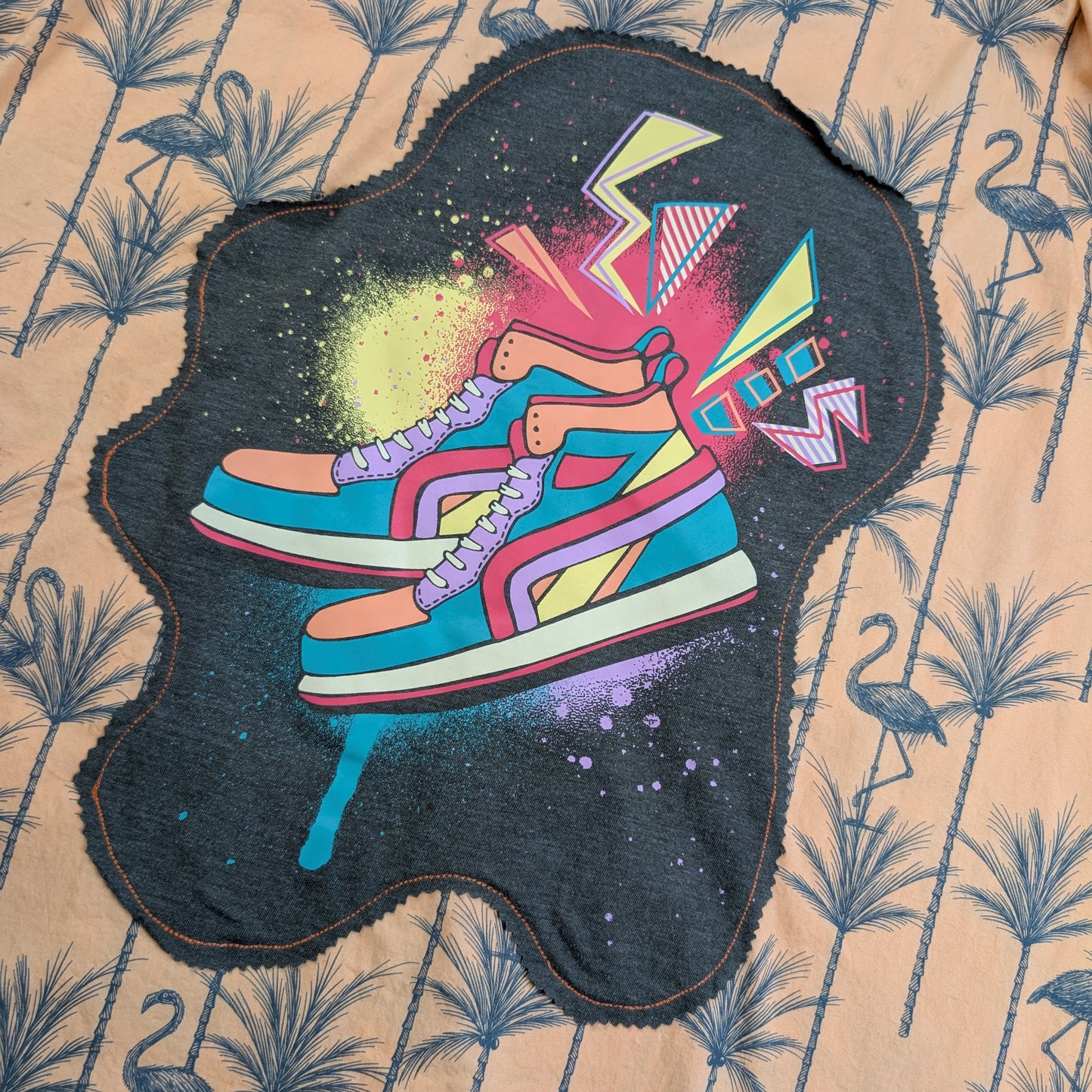 A dark charcoal gray fabric patch featuring a colorful graphic of two sneakers with abstract paint splatters and geometric shapes, laid flat on a beige tropical flamingo-patterned fabric background.
