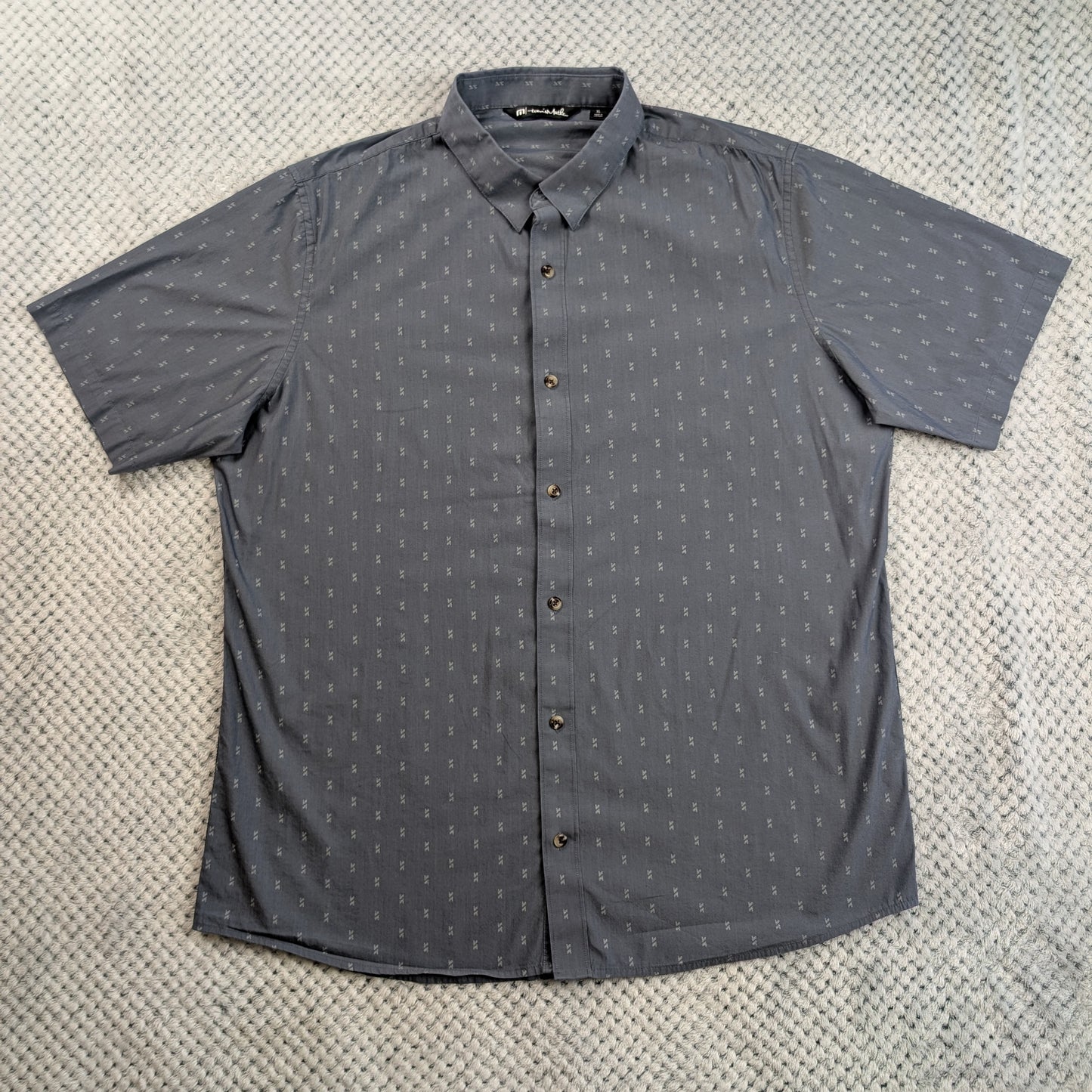 A dark gray short-sleeve button-up shirt with a small repeating white geometric pattern, laid flat on a textured light gray surface.
