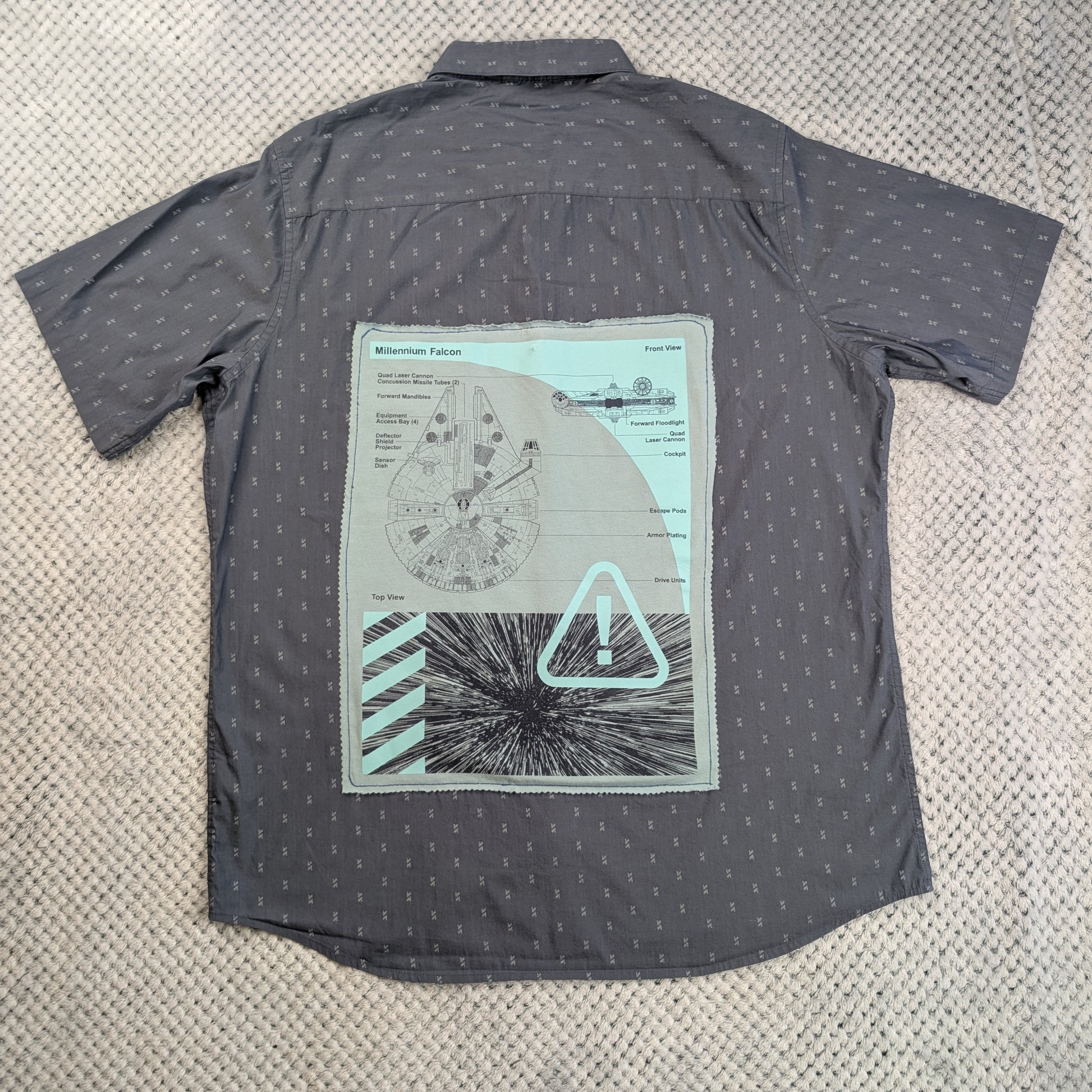A dark gray short-sleeve button-up shirt with a small repeating pattern, laid flat on a textured beige carpet. The back of the shirt features a large graphic print depicting a circular diagram, a warning triangle, and a starburst design in light gray, white, and mint green.