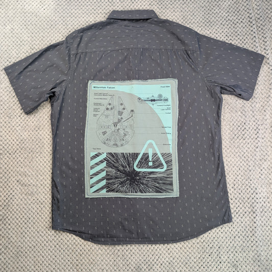 A dark gray short-sleeve button-up shirt with a small repeating pattern, laid flat on a textured beige carpet. The back of the shirt features a large graphic print depicting a circular diagram, a warning triangle, and a starburst design in light gray, white, and mint green.